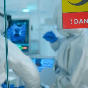 Chemists in coverall trying to develop vaccine using tablet standing behind the glass wall working in danger area of lab. Scientists examining virus evolution researching for treatment against covid19